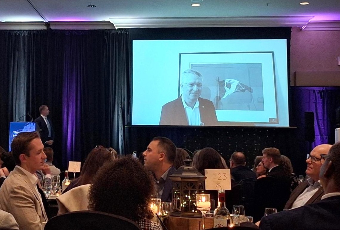 Audience seated at a formal dinner event watching a speaker projected on a large screen on stage.