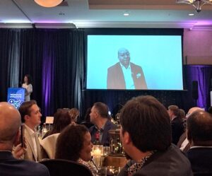 Audience seated at a formal dinner event watching a speaker projected on a large screen on stage.