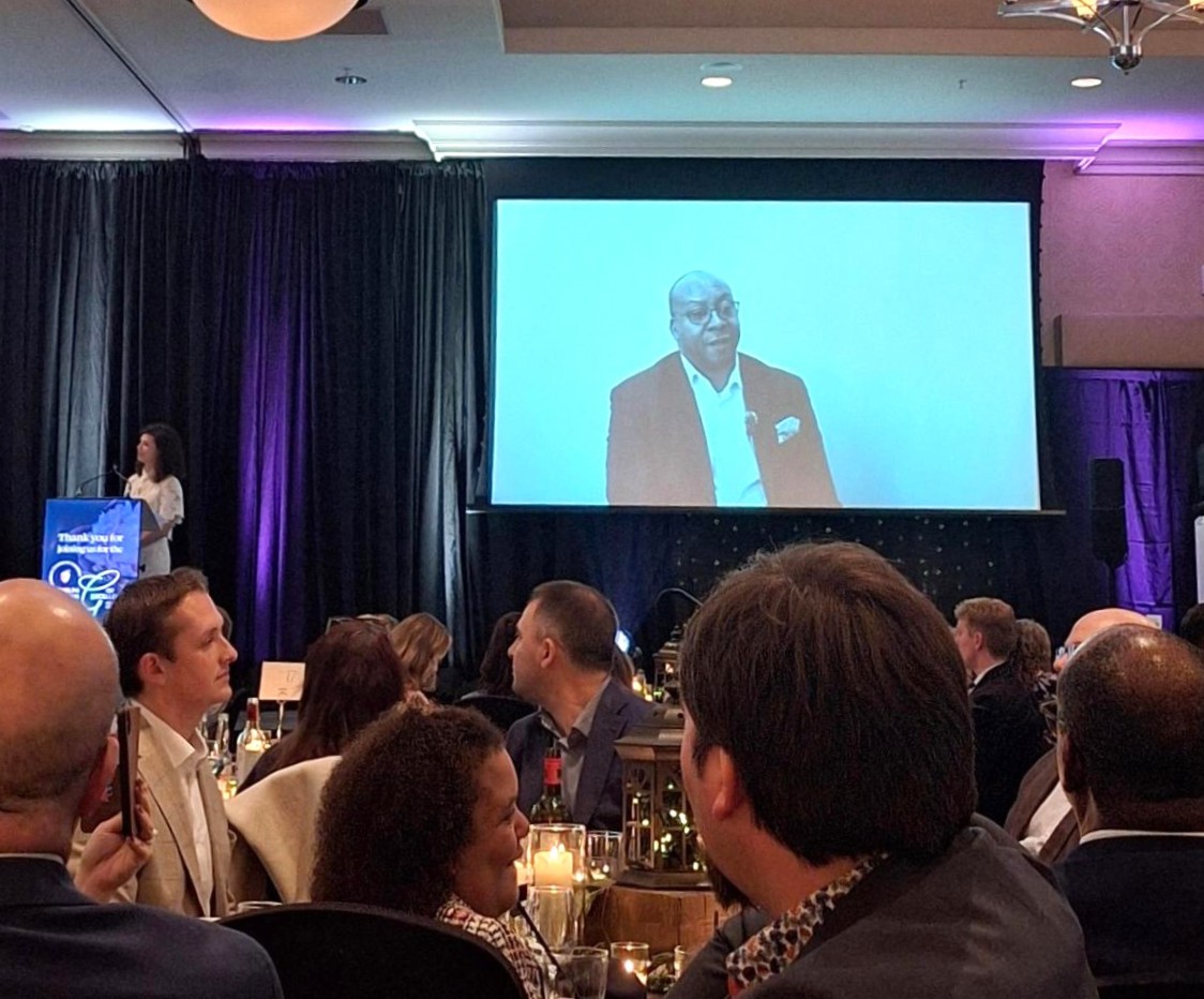 Audience seated at a formal dinner event watching a speaker projected on a large screen on stage.