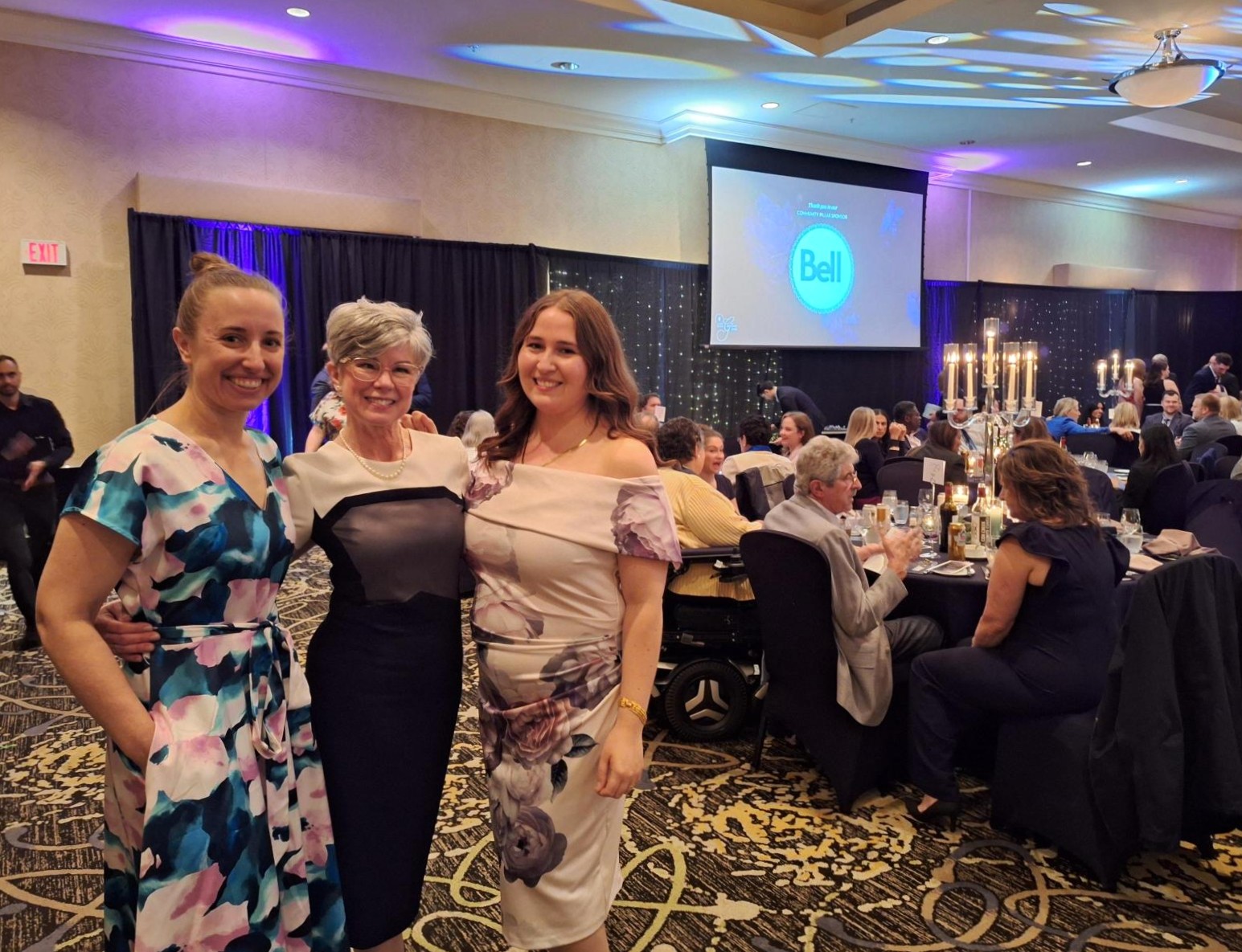 Three women in elegant dresses posing together at a formal banquet with seated guests
