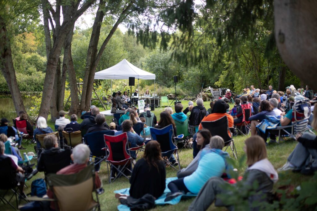 Outdoor gathering with people seated on folding chairs listening to speakers under a white canopy in a wooded park.