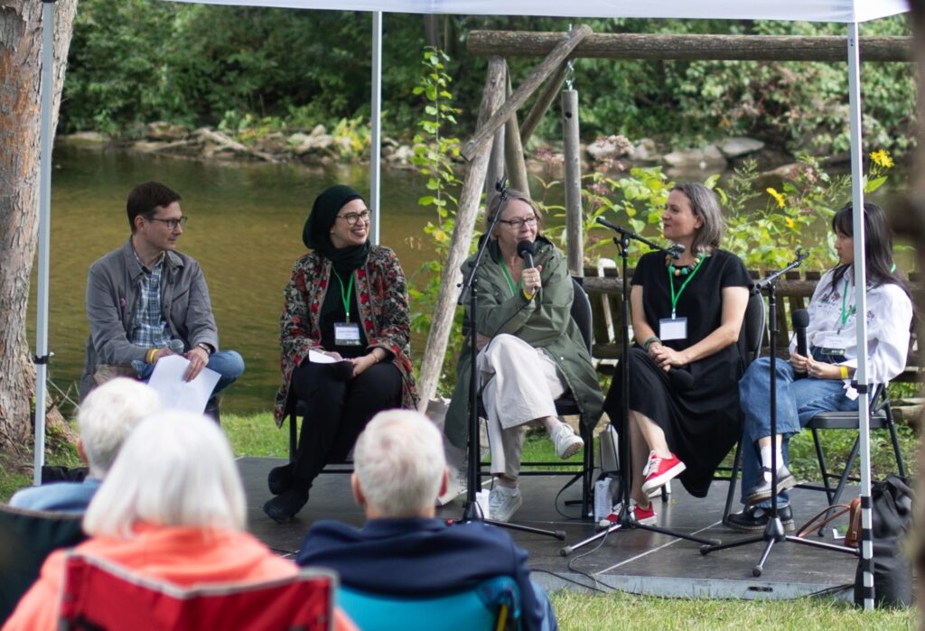 Panel of five speakers seated outdoors under a canopy near a river, engaging with an audience during a discussion event.