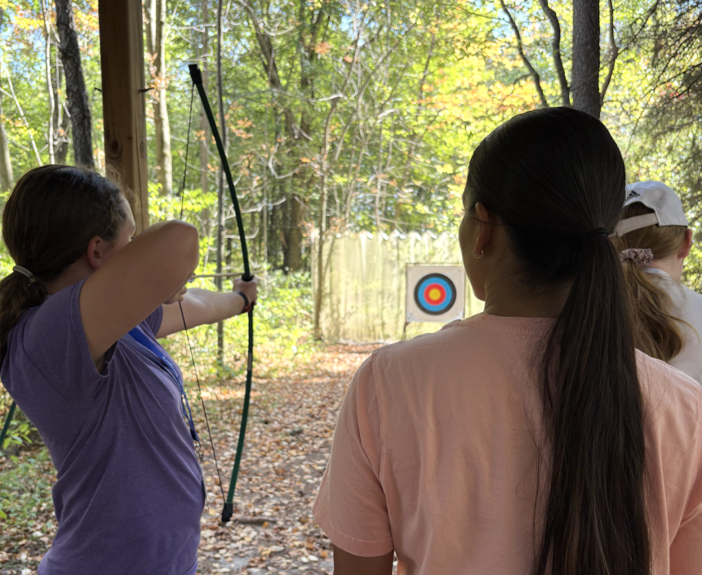 Children engage in archery on a forest path, focusing on hitting a multicolored target placed ahead.