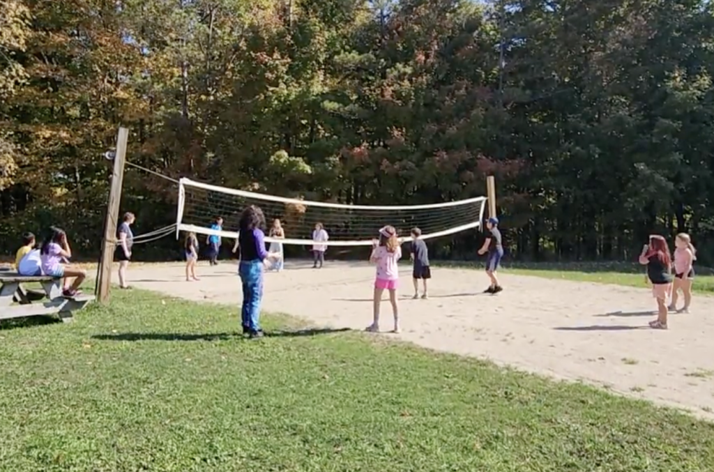 Children playing volleyball on a sandy court surrounded by green grass and trees in a sunny outdoor setting.