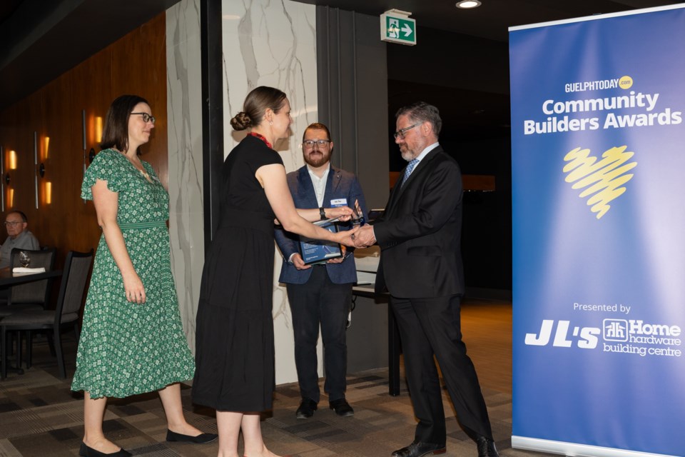 Two women and two men at an indoor awards ceremony, one woman shaking hands with a man while receiving an award plaque near a Community Builders Awards banner.