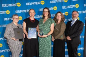 Five individuals stand in front of a blue GuelphToday.com backdrop, three women holding awards and two others posing formally.