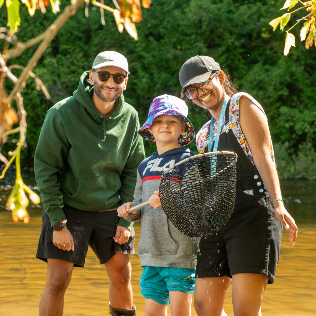 Two adults and a child posing near a calm river, the child gripping a fishing net with greenery in the background.