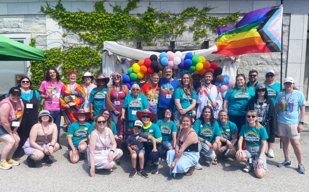 Community members celebrating pride day, standing and sitting together under a vibrant balloon arch and a pride flag.