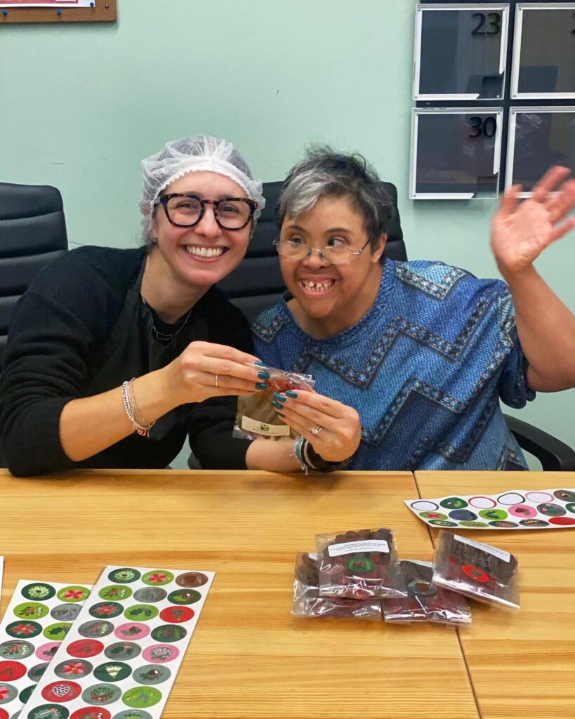Two women smiling and holding packaged treats at a table with colorful stickers and more packaged items in front of them.