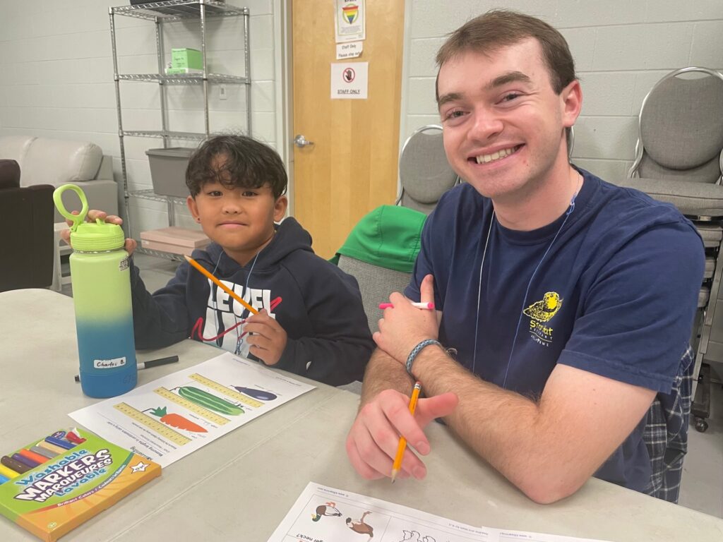 Young boy and adult male sitting at a table working on colorful worksheets with pencils and markers in a classroom setting.