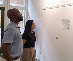 Two people attentively reading a small text display on a white wall inside a bright room with large windows.