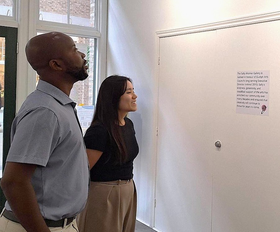 Two people attentively reading a small text display on a white wall inside a bright room with large windows.