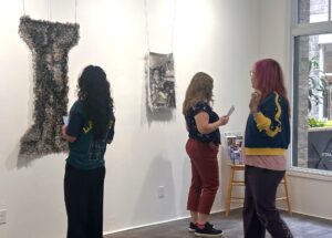 Three women observe textured fiber art pieces displayed on a white gallery wall in a bright room with large windows.