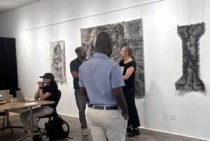Three people engage in conversation in an art gallery with textured abstract wall hangings displayed behind them.