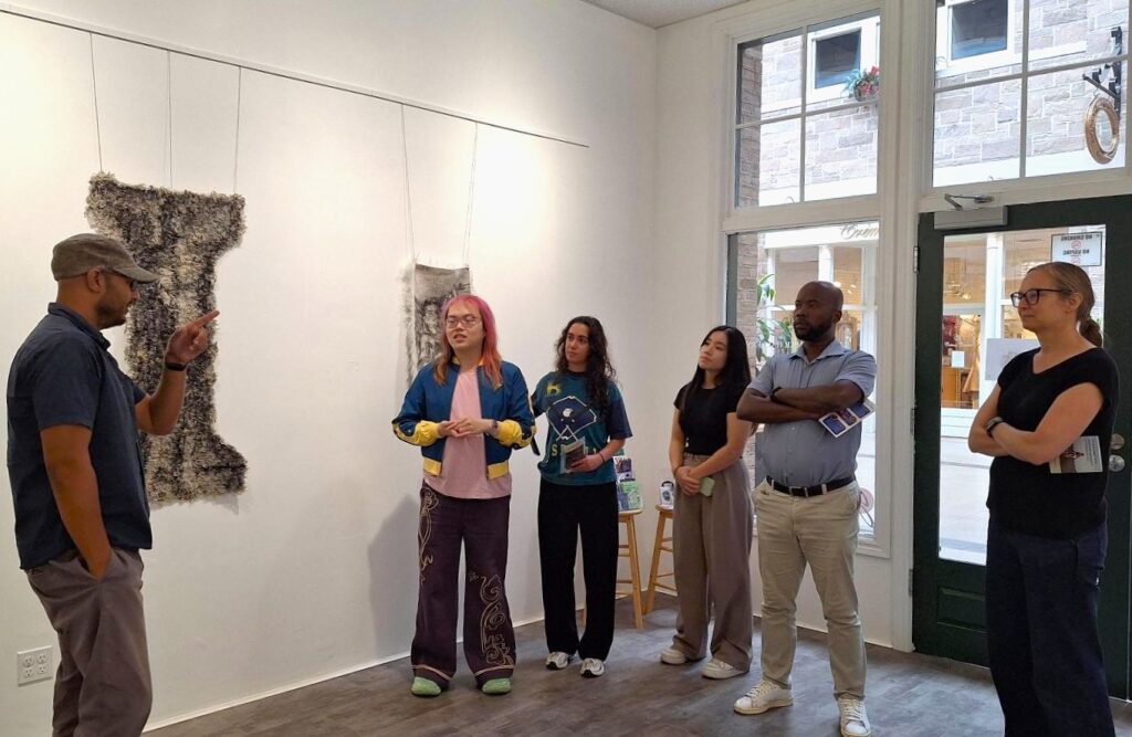 A man wearing a cap explains artwork on a gallery wall to a diverse group of five attentive people standing in a bright gallery room with large windows.