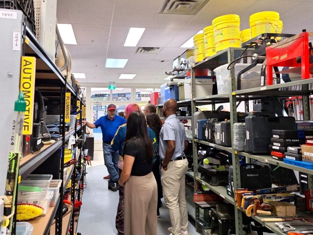 Group of people listening to a man explaining tools in a well-organized hardware store aisle.