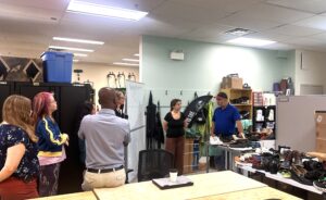 A group of people attentively listening to a man explaining shoe designs in a workshop filled with footwear and storage units.