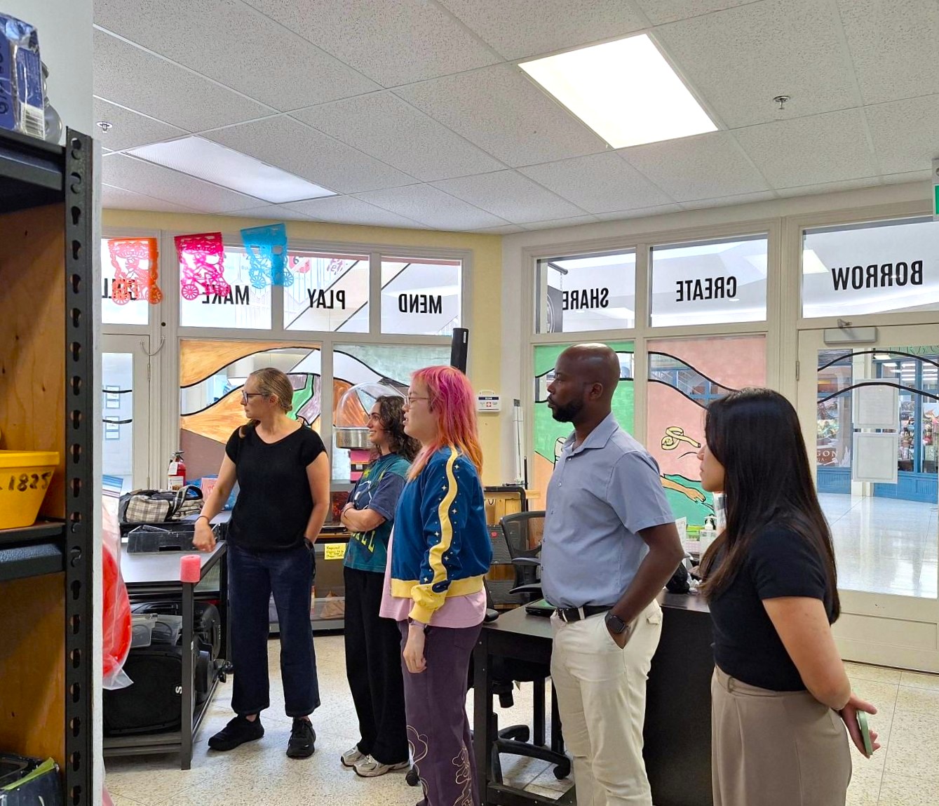 A group of five diverse adults attentively listening looking to the left inside a bright, colorful area with words on the windows.