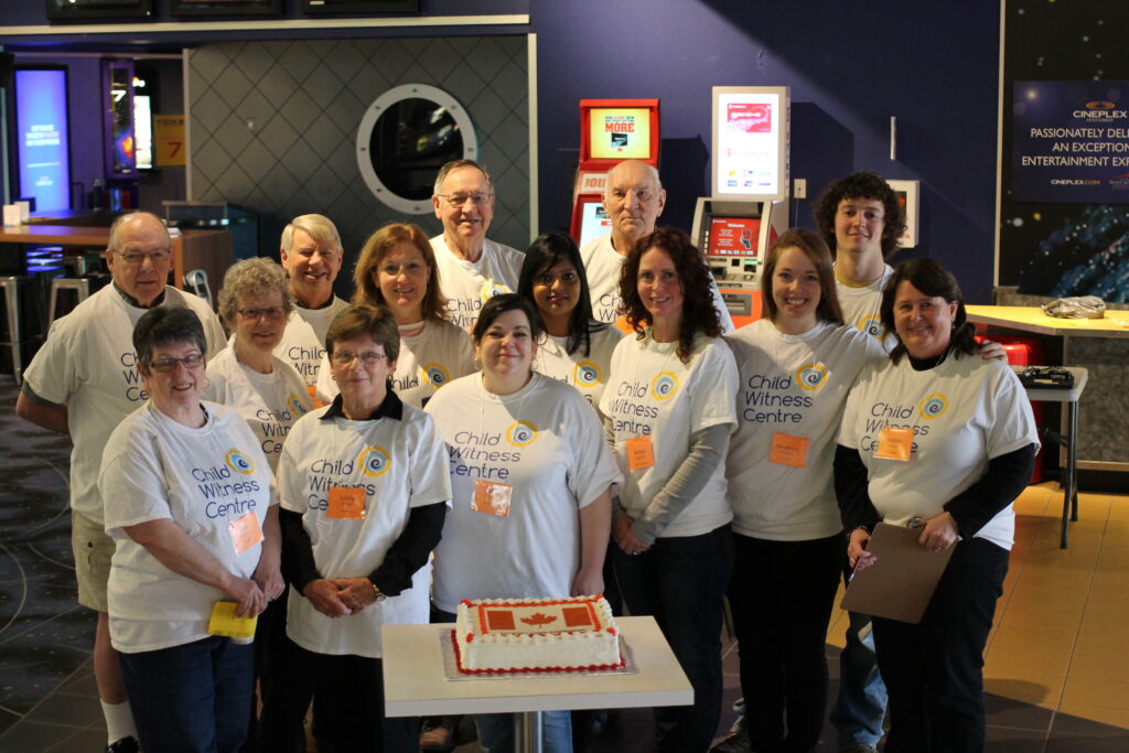 Group of volunteers wearing Child Witness Centre t-shirts gathered around a table with a decorated cake indoors.