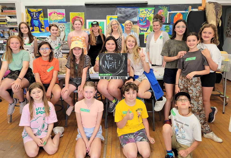 Group of diverse children and adults smiling in an art classroom with colorful paintings on the walls.