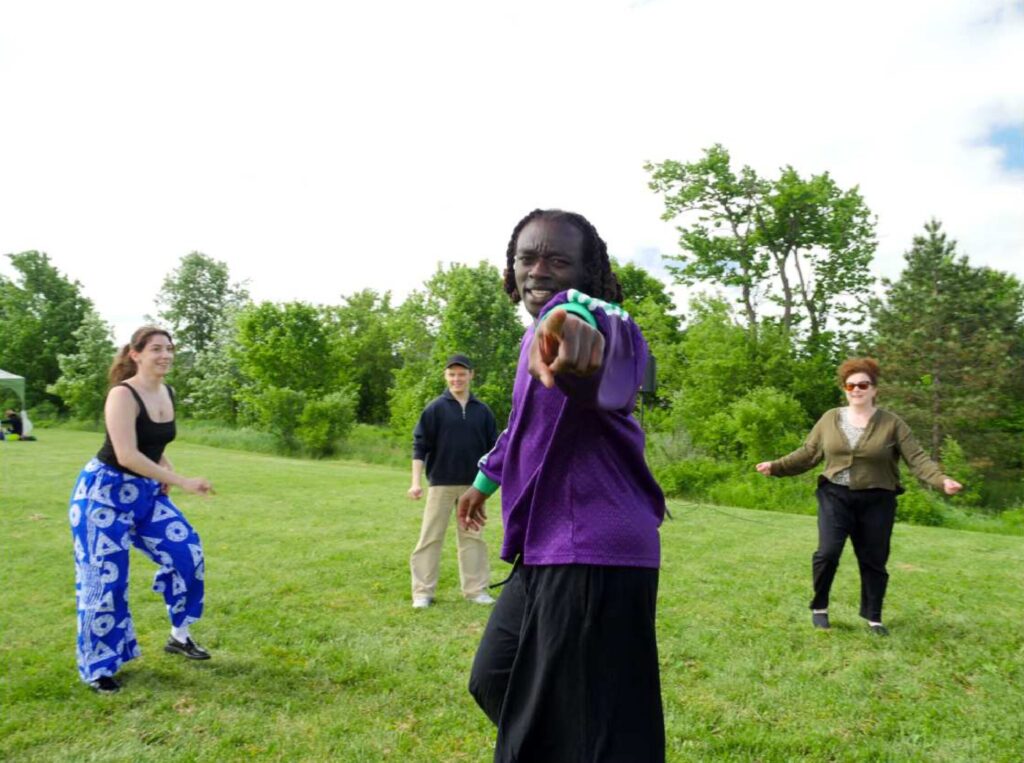 Four people dancing on a sunny day outdoors, standing on green grass with trees in the background, one man pointing playfully at the camera.