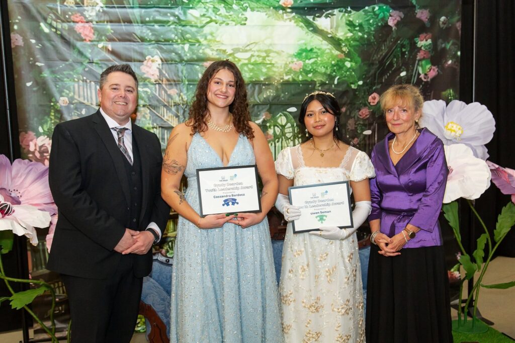 Two young women in formal dresses holding certificates, standing between a man in a suit and a woman in a purple blouse, with a floral backdrop.