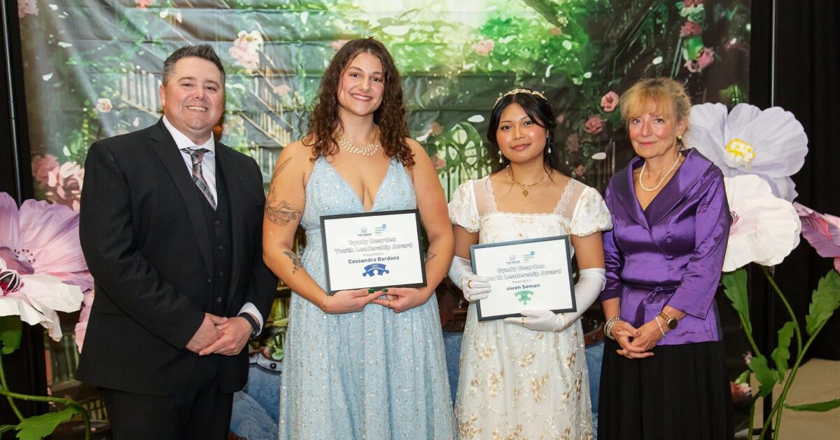 Two young women in formal dresses holding certificates, standing between a man in a suit and a woman in a purple blouse, with a floral backdrop.