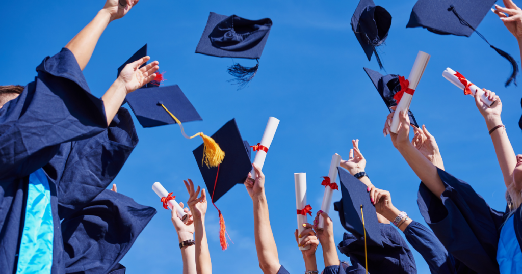 Graduates in navy gowns toss caps and hold diplomas high against a clear blue sky in celebration.