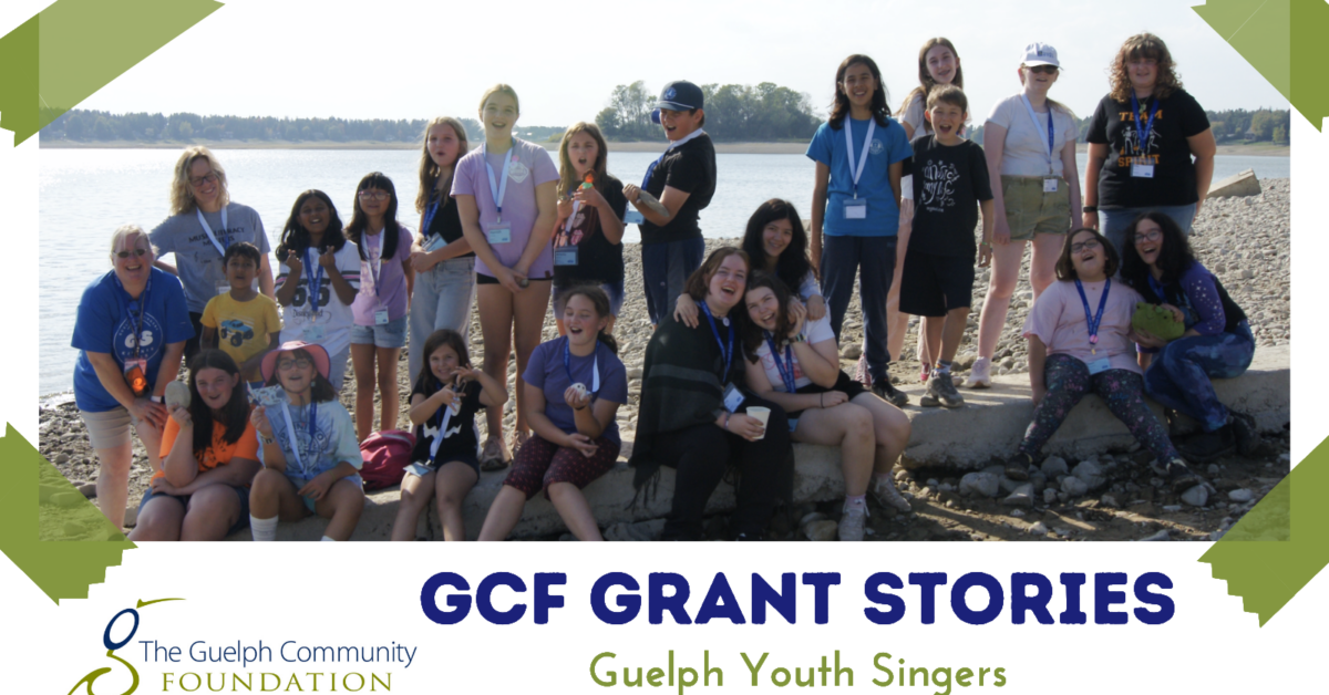 Group of diverse youth singers posing happily on a rocky shore by the water under clear skies.