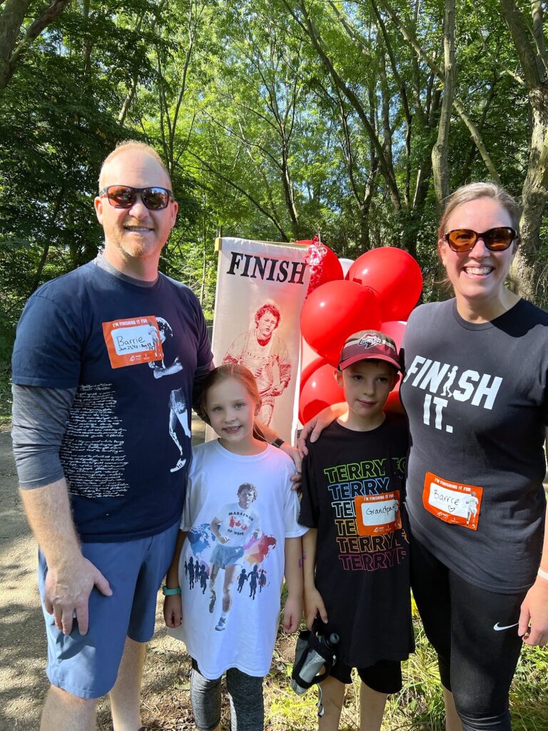 Smiling family of four posing outdoors near a finish line banner and red balloons during a community event.