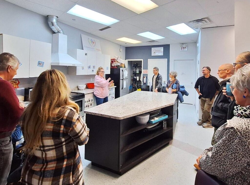 Group of adults attentively listening to a woman speaking in a modern kitchen with a large island and white cabinets.