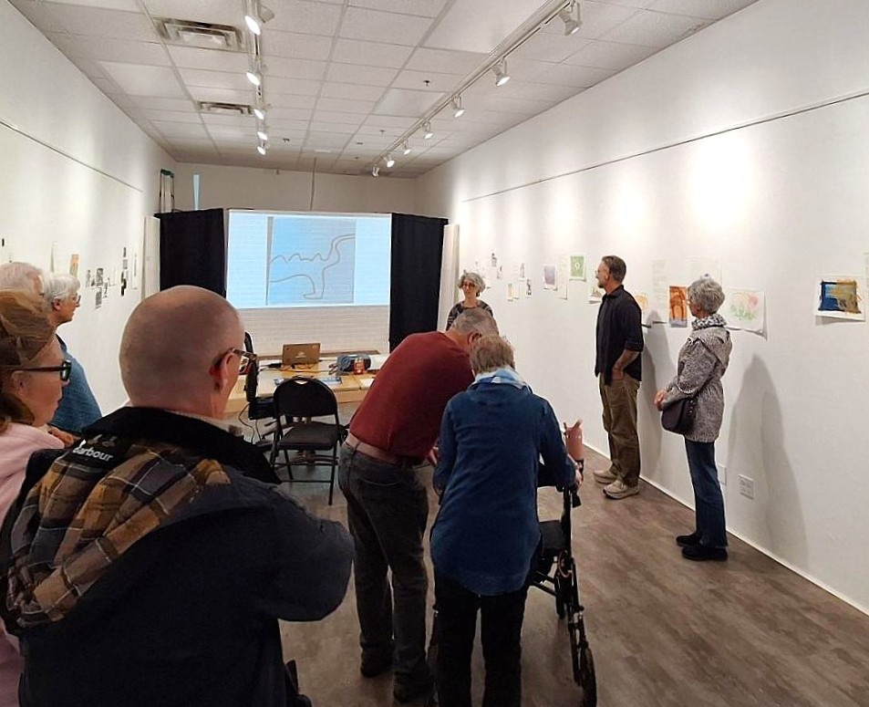 Group of people attentively viewing an art presentation in a gallery with drawings displayed on white walls.
