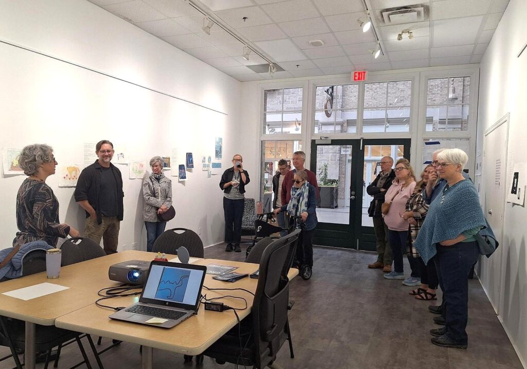 A group of people attentively listening to a speaker inside a bright art gallery with white walls and displayed artworks.