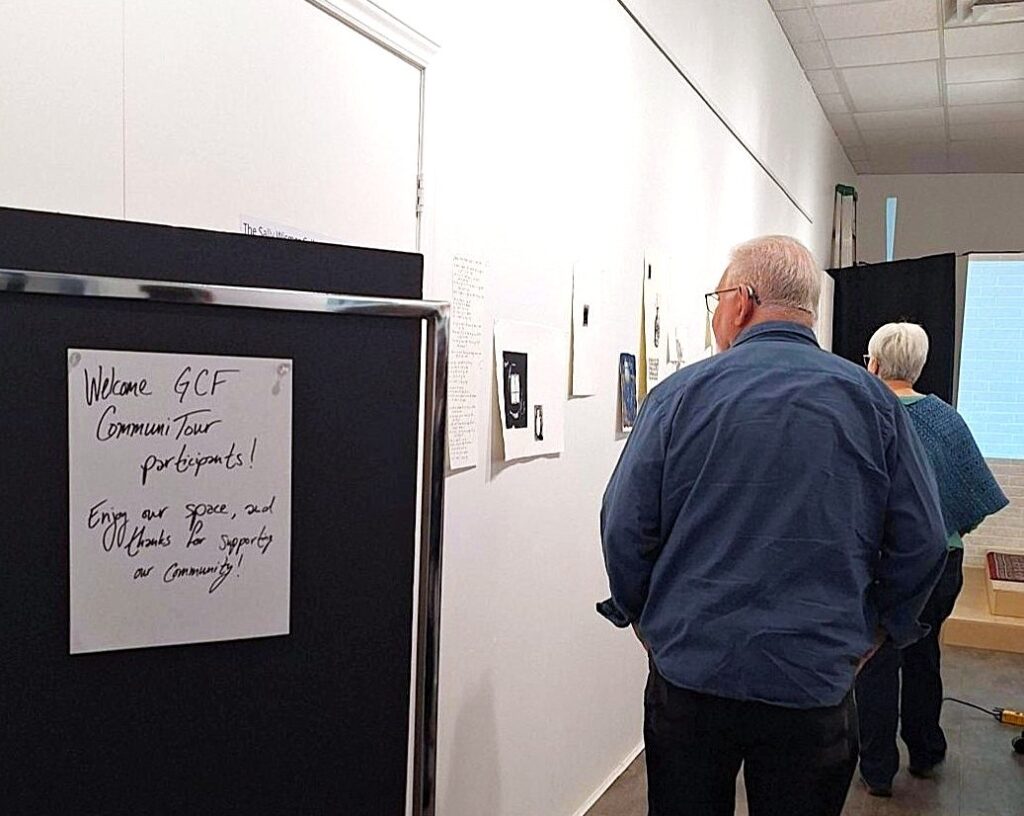 Two older adults view artwork displayed on a white gallery wall. Sign that reads "Welcome GCF CommuniTour".