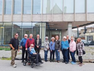 Group of twelve adults posing outdoors in front of a metal fence and building, dressed casually for cool weather.