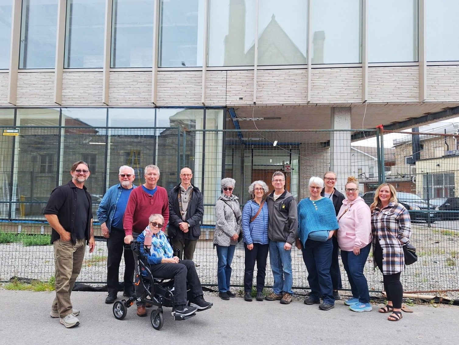 Group of twelve adults posing outdoors in front of a metal fence and building, dressed casually for cool weather.