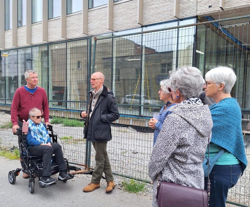 Group of older adults gathered outdoors near a fenced construction site, engaged in conversation.