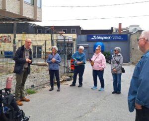 A man speaks to a small group of adults standing outdoors near a fenced construction area in an urban setting.