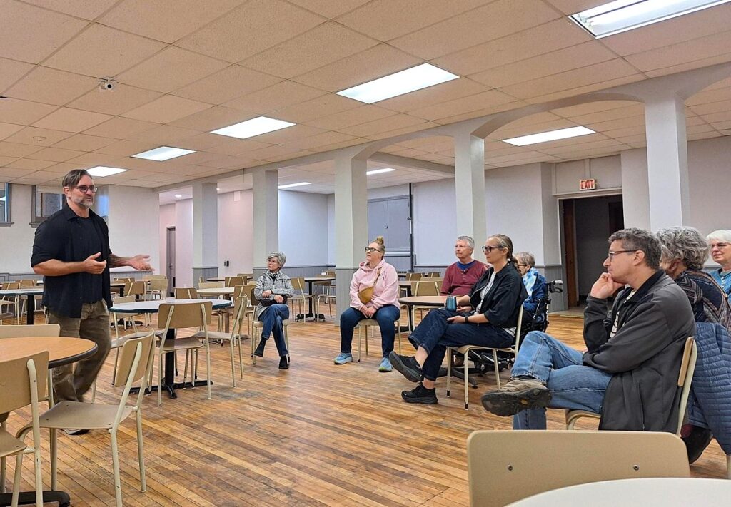 Man standing and speaking to a small group seated in a spacious church basement room with wooden floors.