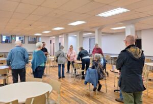 A group of people socializing and standing in a spacious basement church room with wooden floors