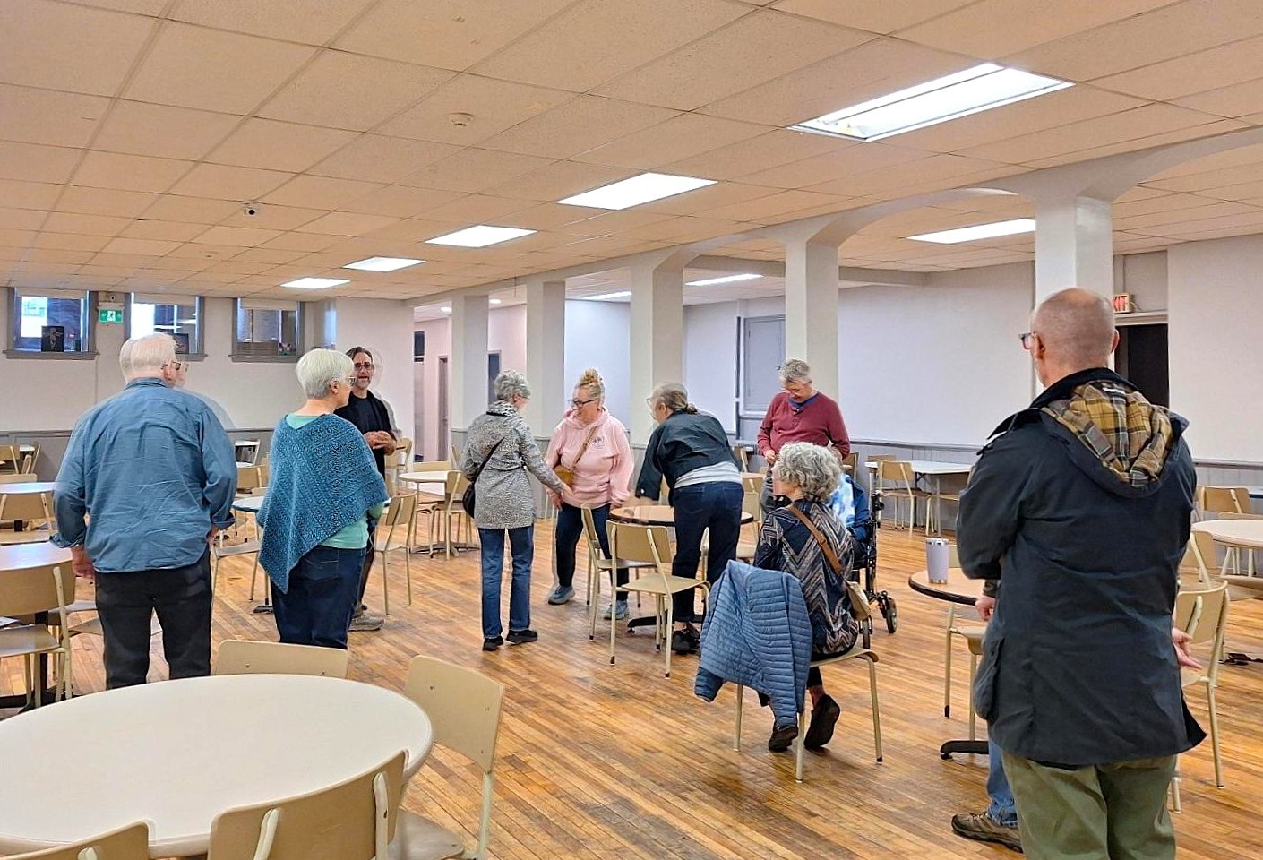 A group of people socializing and standing in a spacious basement church room with wooden floors