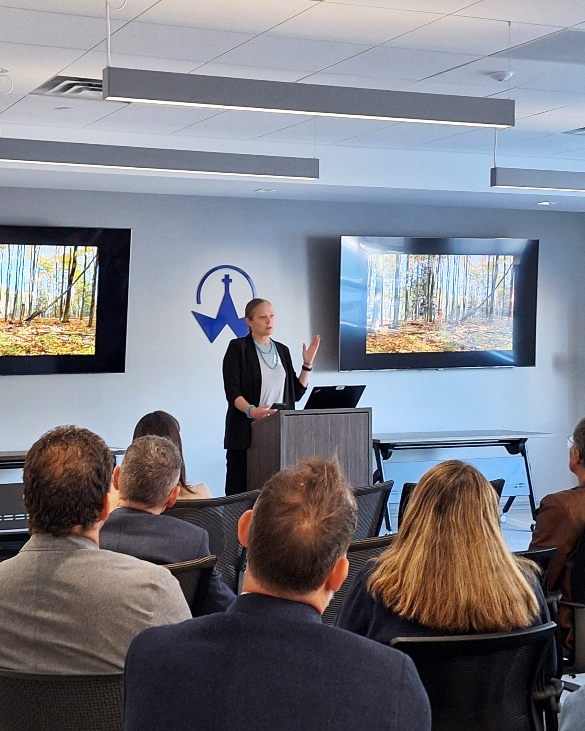 A presenter (Sarah Haanstra) speaks from a podium to an audience, with visuals of a forest displayed on screens behind the speaker.