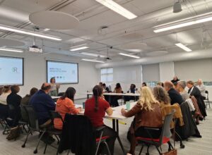 A presenter leads a workshop session while attendees seated around tables listen and participate in discussion during the Finding Abundance symposium.