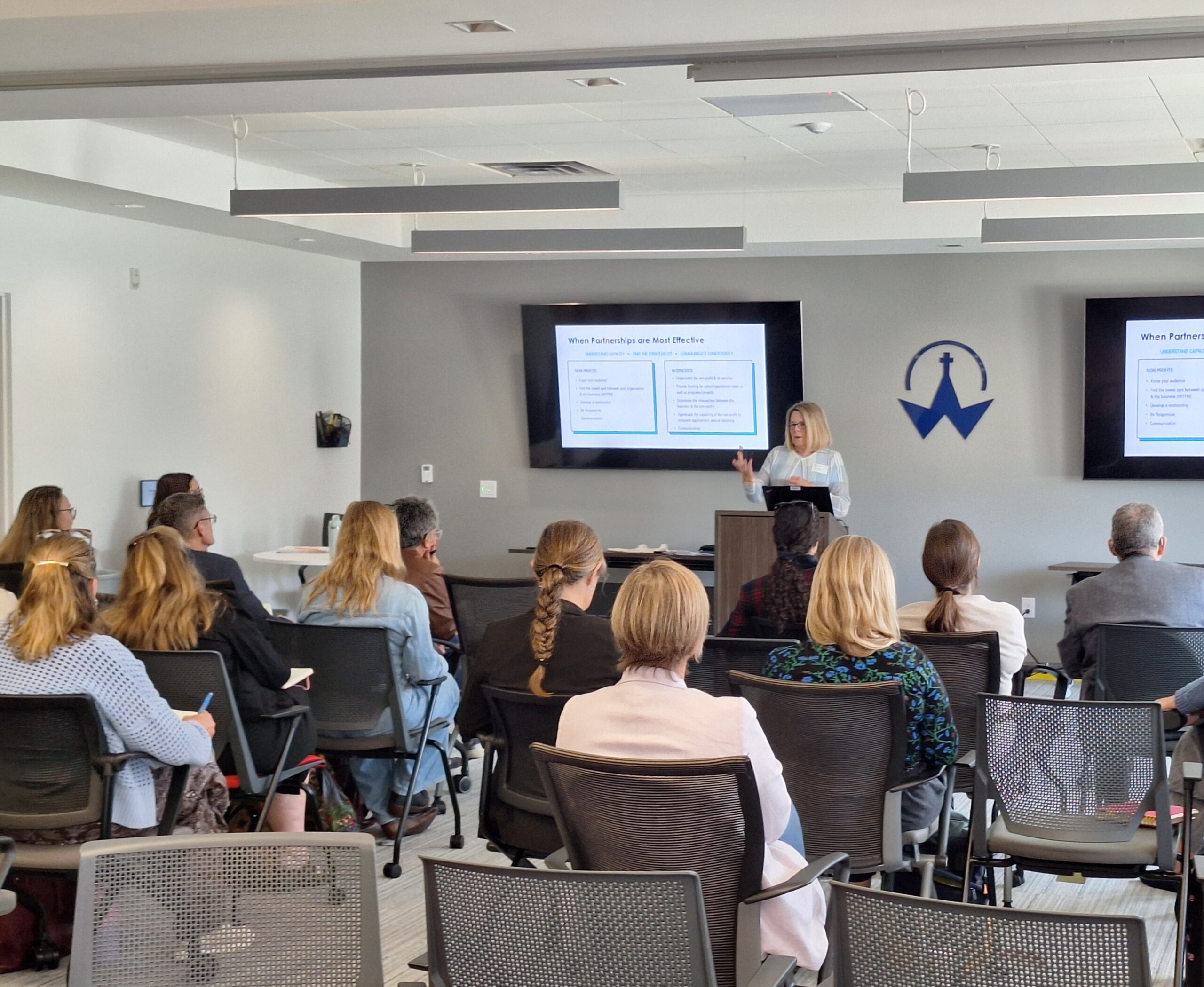 A speaker presents to attendees during a workshop on building effective partnerships, with presentation slides displayed on screens at the front of the room.
