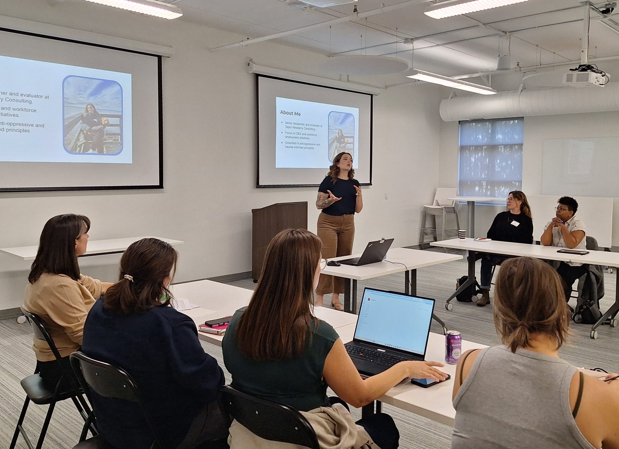 A workshop facilitator presents to attendees seated around tables during a session, with presentation slides displayed at the front of the room.
