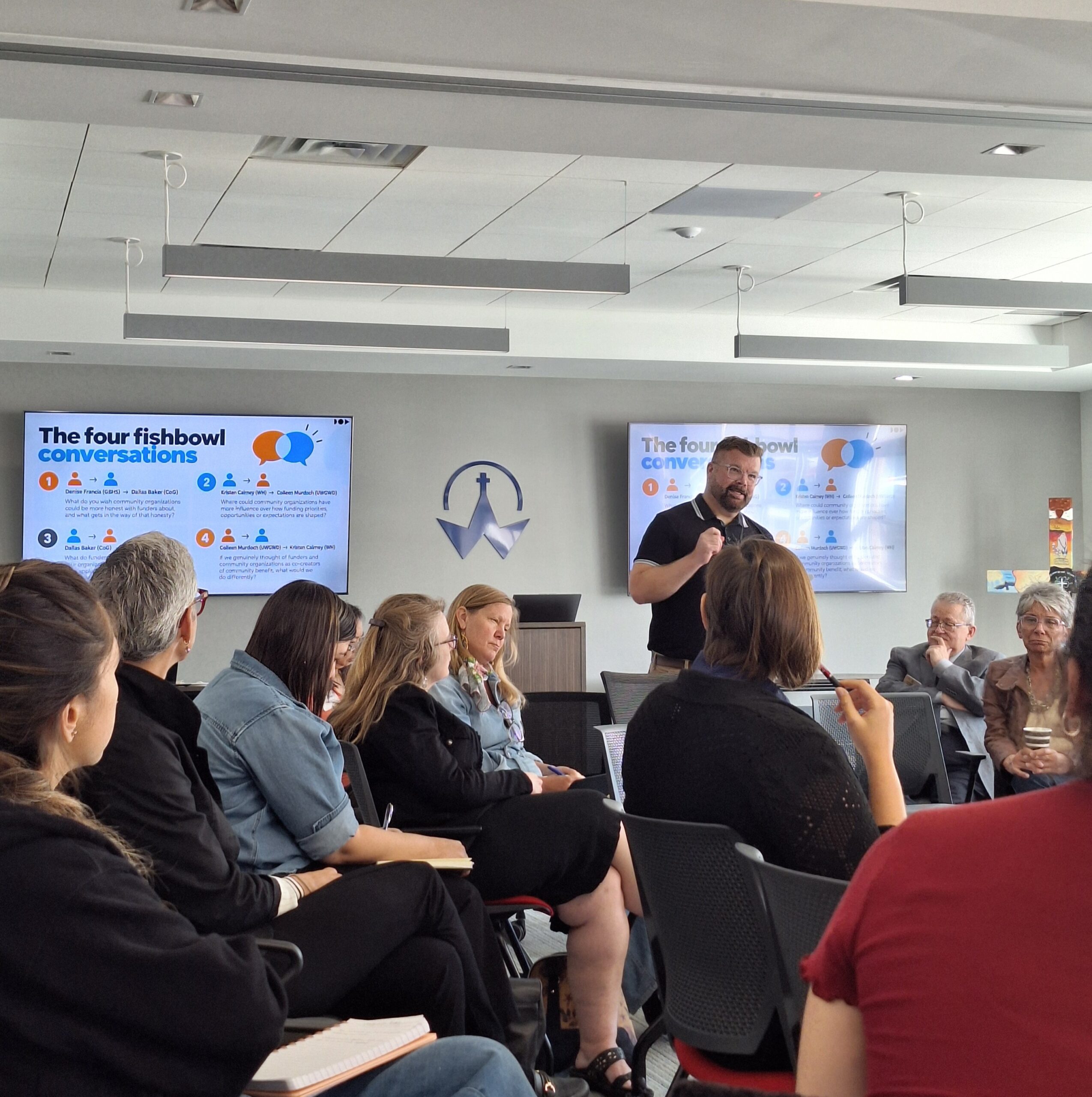 A workshop facilitator presents to attendees seated in circles during a session, with presentation slides displayed at the front of the room.
