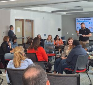 A workshop facilitator presents to attendees seated in circles during a session, with presentation slides displayed at the front of the room.