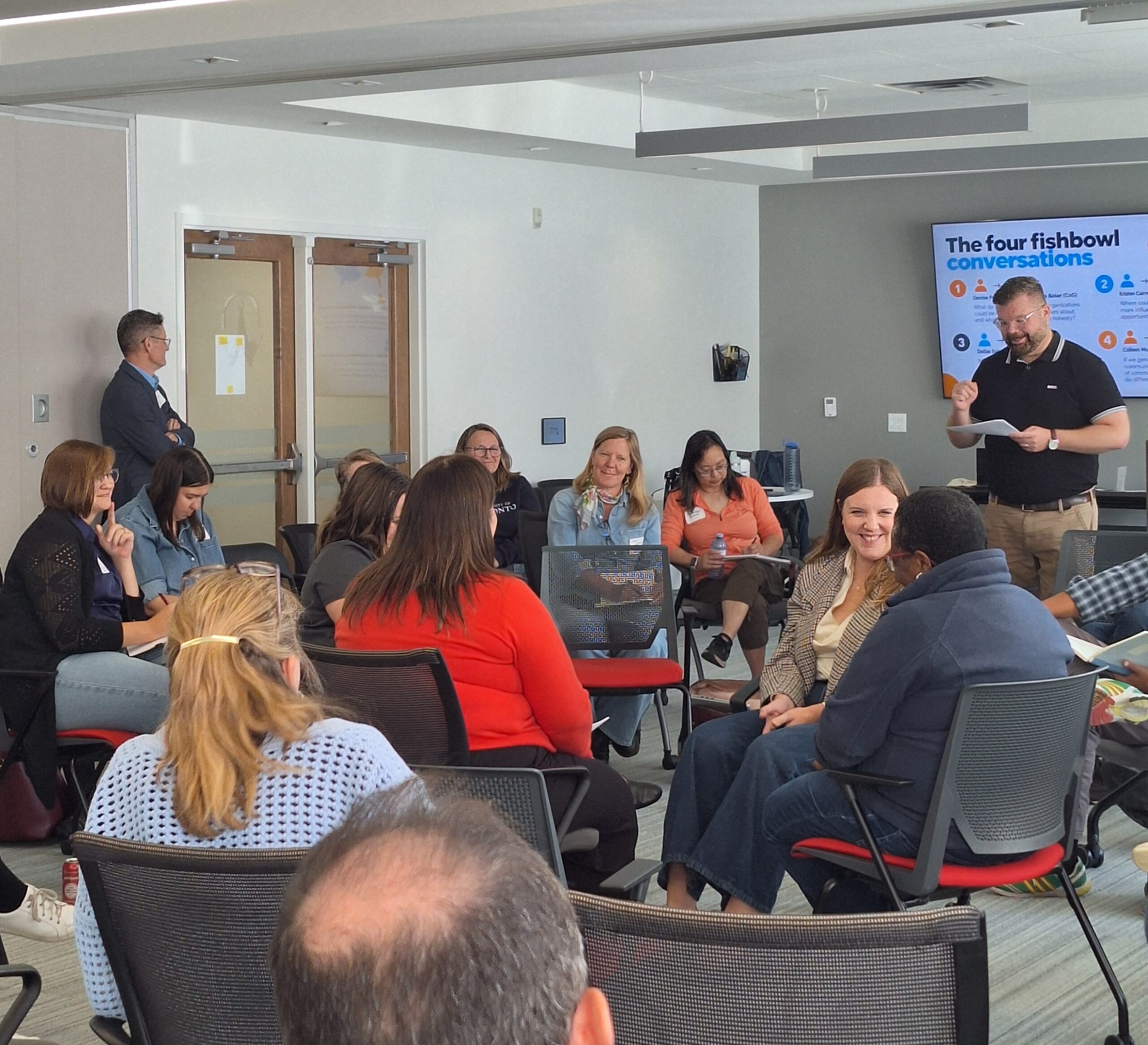 A workshop facilitator presents to attendees seated in circles during a session, with presentation slides displayed at the front of the room.