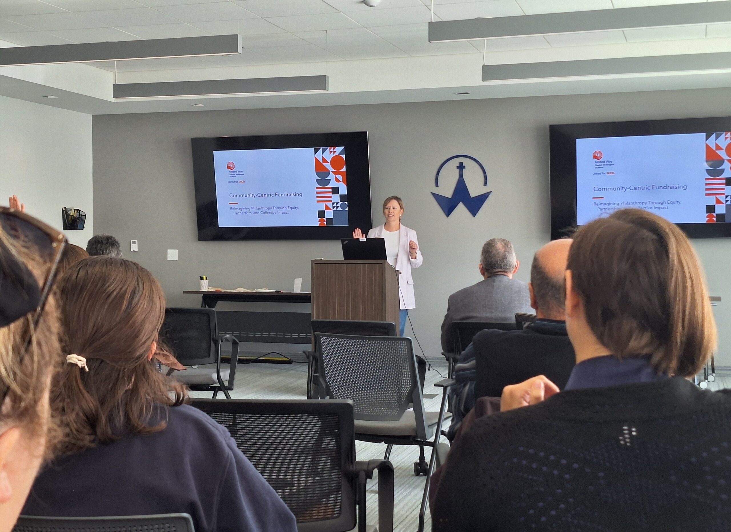 A presenter speaks to attendees during a workshop on community-centric fundraising, with presentation slides displayed on screens at the front of the room.