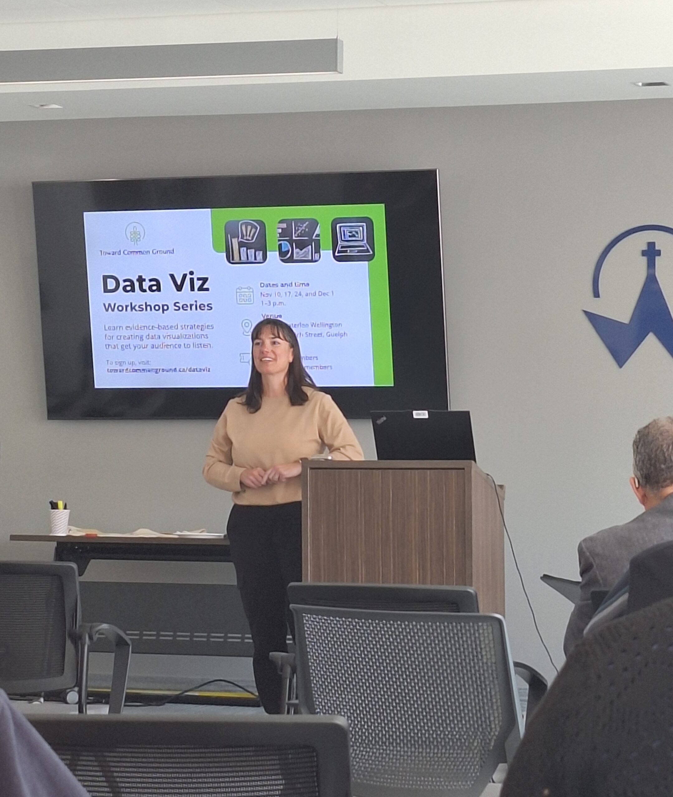 A presenter speaks at the front of the room while attendees listen from the audience.